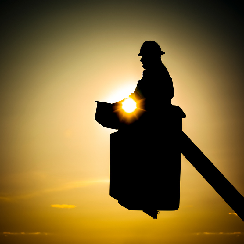 A lineman in a cherry picker backlit by the sunset